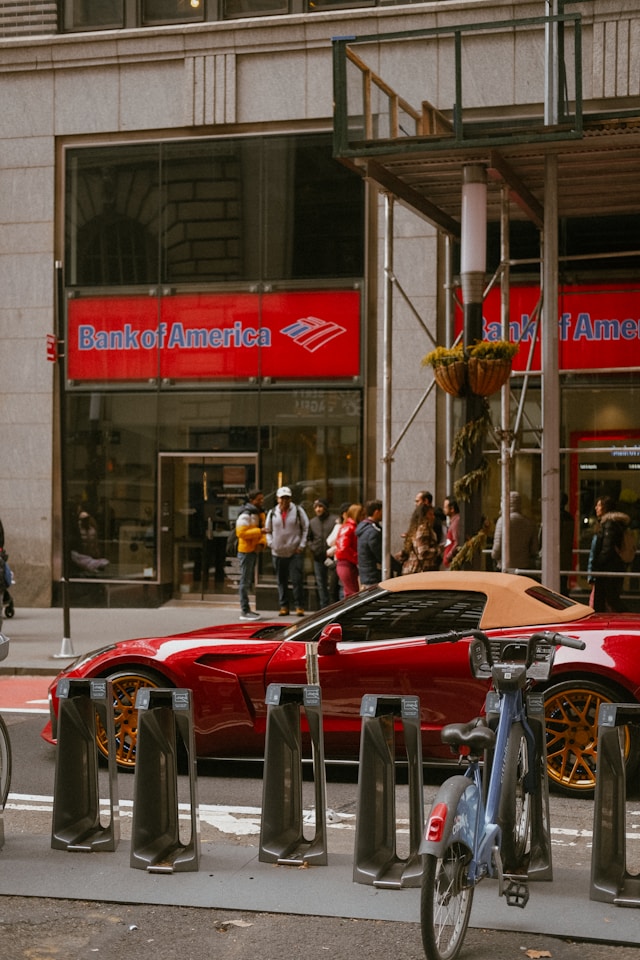 A red sports car next to a Bank of America building.