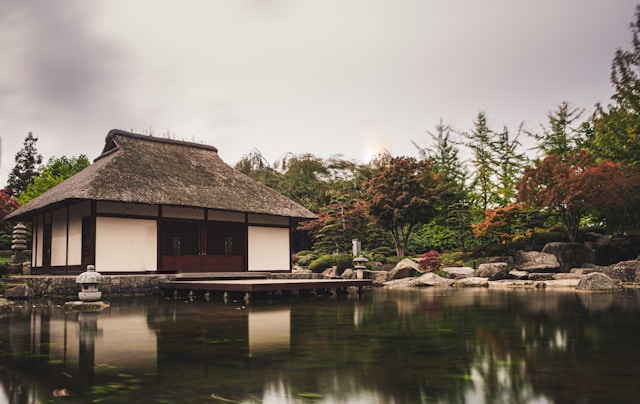 A brown Japanese house next to a body of water, surrounded by trees.