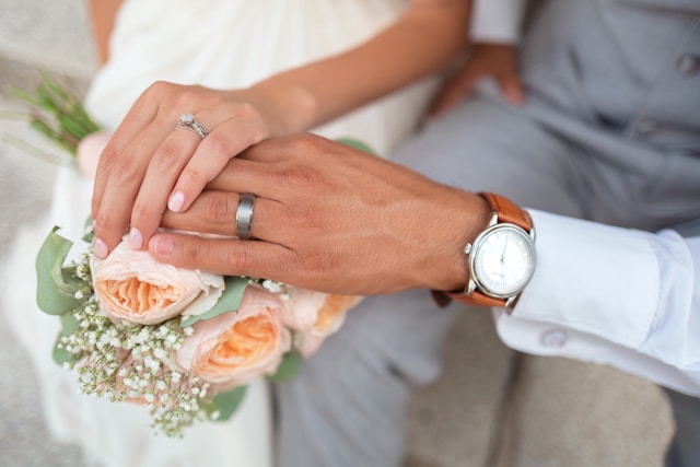 A close-up of a bride and bridegroom holding their hands above a bouquet of pink roses.