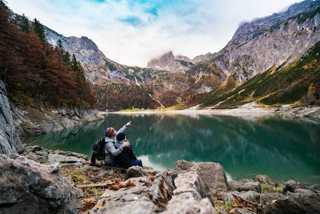 Two people sit on a wooden plank by a lake surrounded by mountains.
