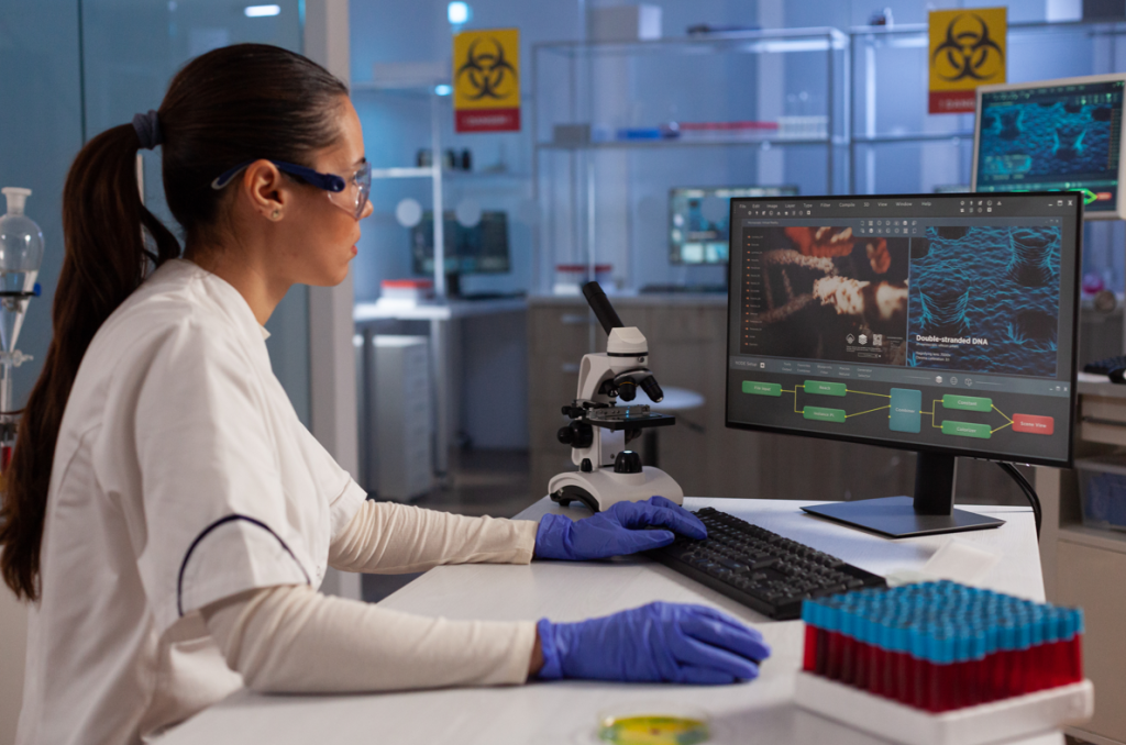 A scientist in a lab coat and gloves is analyzing something on a computer screen beside a microscope and test tubes.