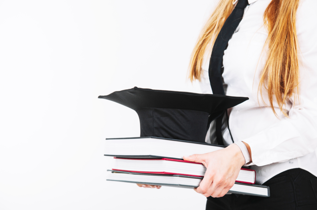 A student in formal attire holding books with a graduation cap on top.