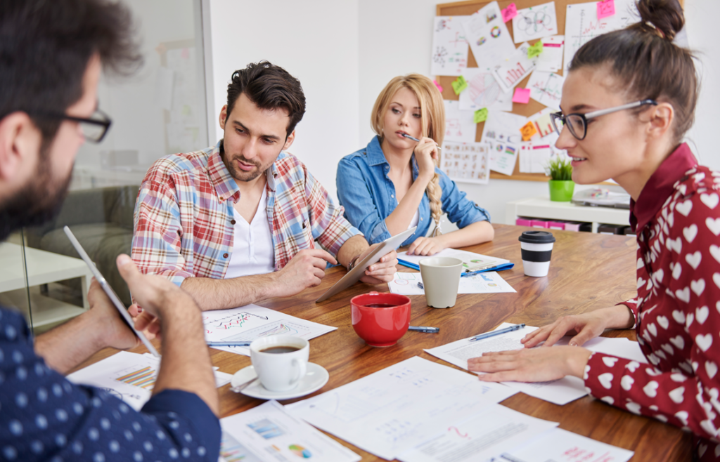 A group of four people having a discussion around a table with charts, documents, and coffee cups.